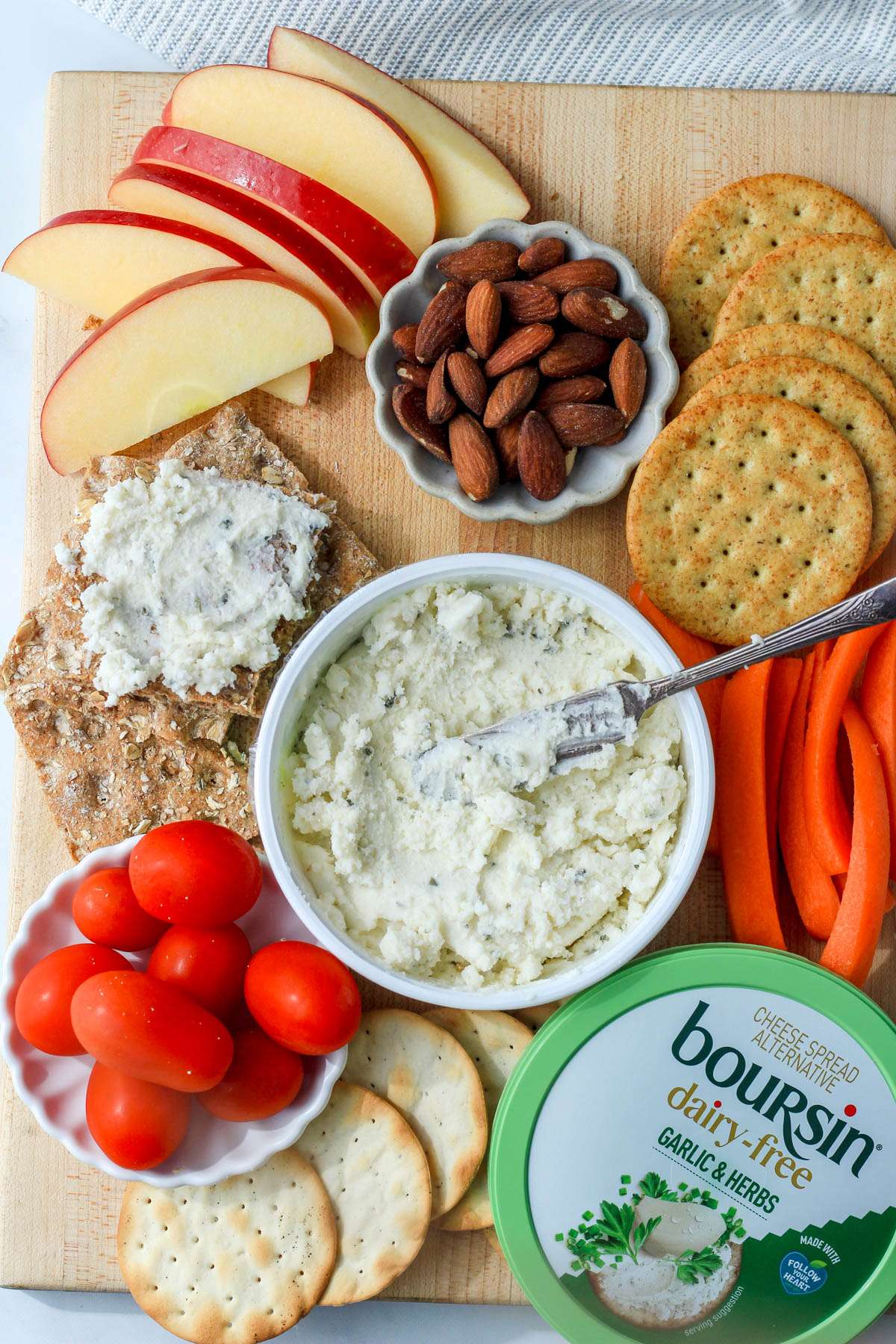 A wooden cutting board topped with dairy-free boursin, crackers, fruit and nuts with a small lid in the bottom right.