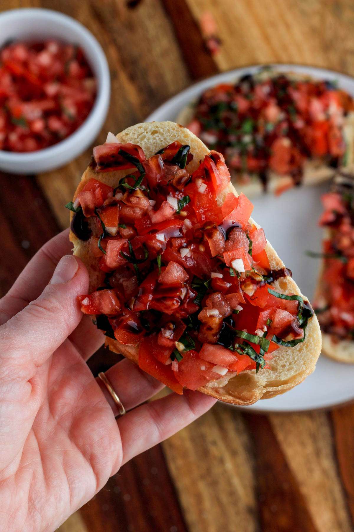 A hand holding a piece of vegan bruschetta toast topped with balsamic glaze over a white plate of toast.