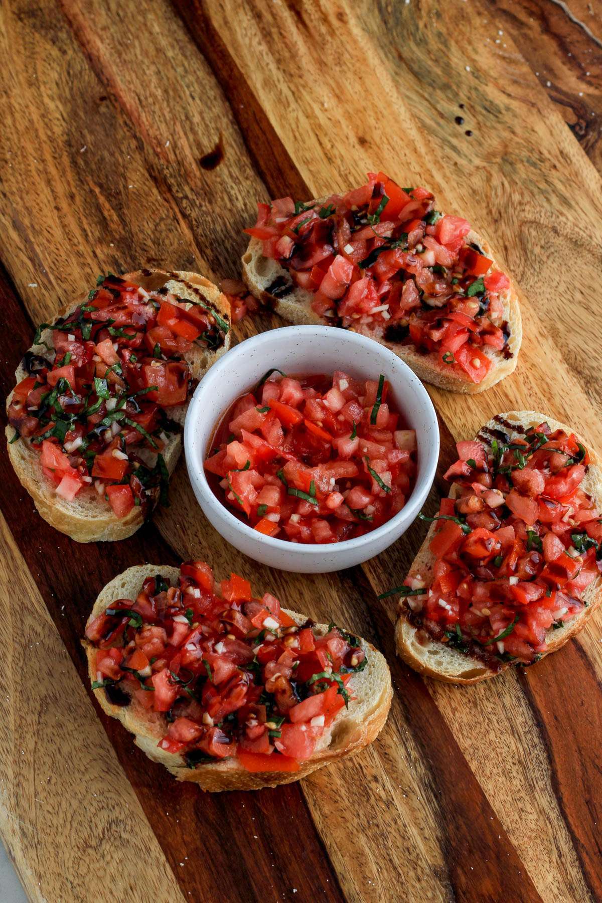 A small white bowl of vegan bruschetta topping surrounded by four pieces of bruschetta toast on a wooden counter.