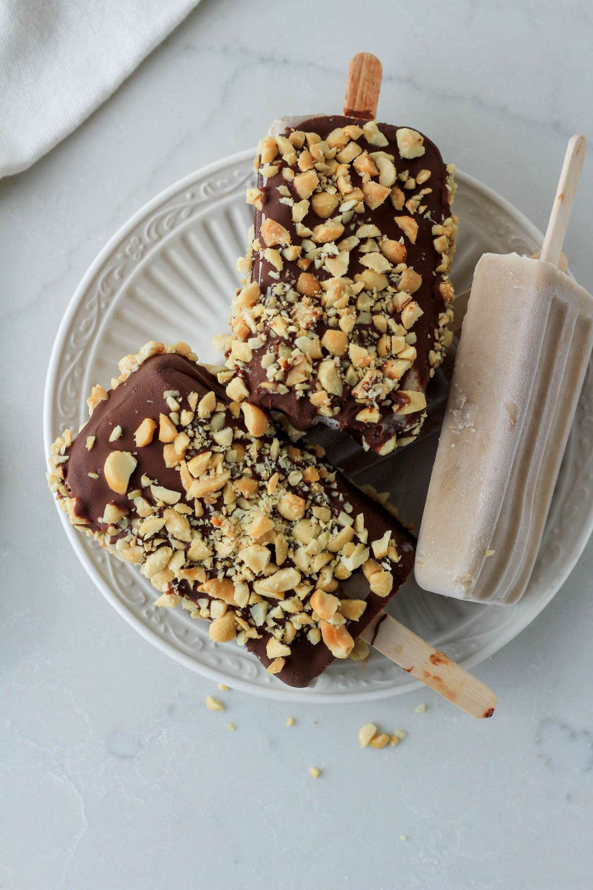 A top down picture of three peanut butter banana popsicles two dipped in chocolate on a white plate.