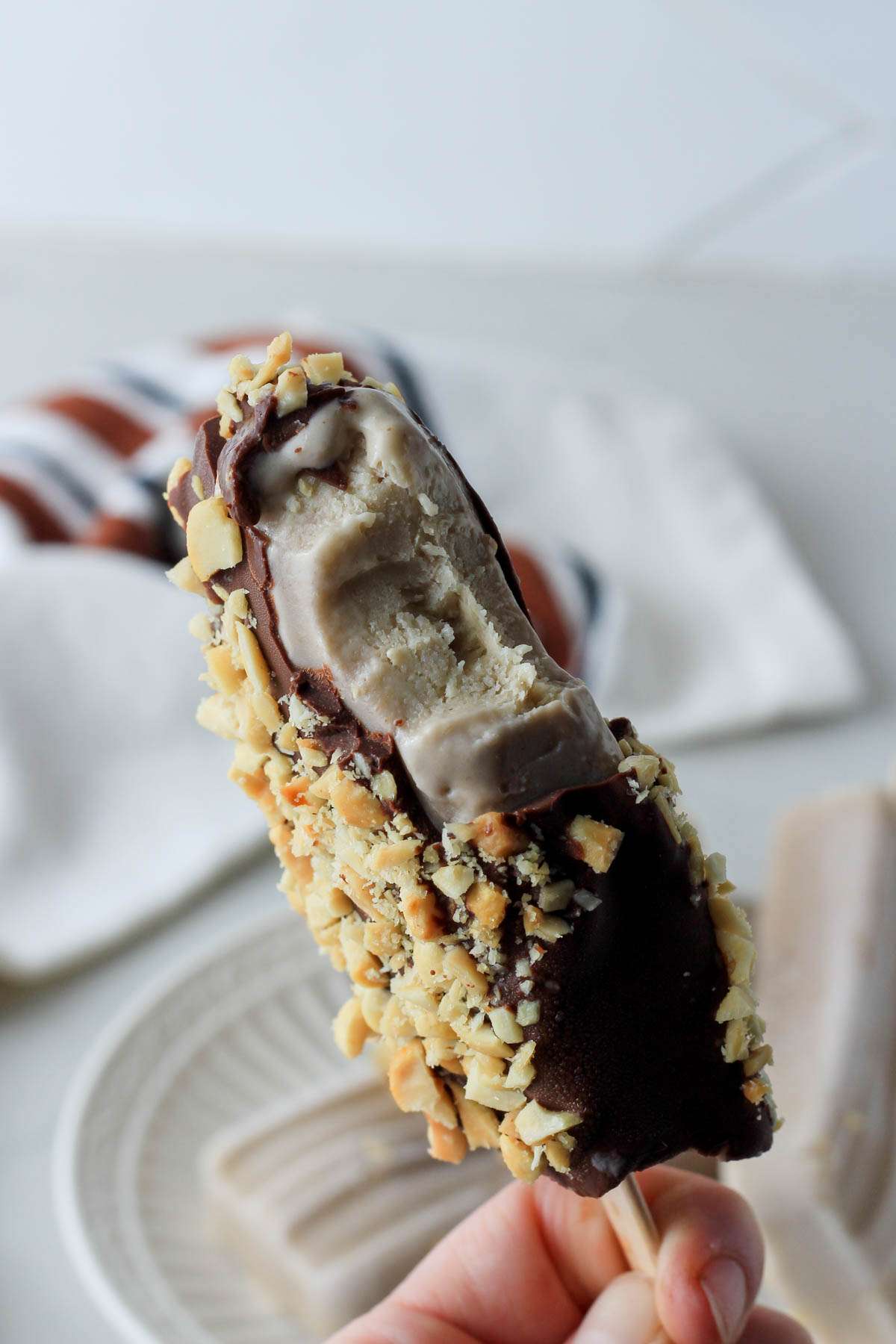 A hand holding a peanut butter banana popsicle dipped in chocolate and peanuts with a bite taken out.