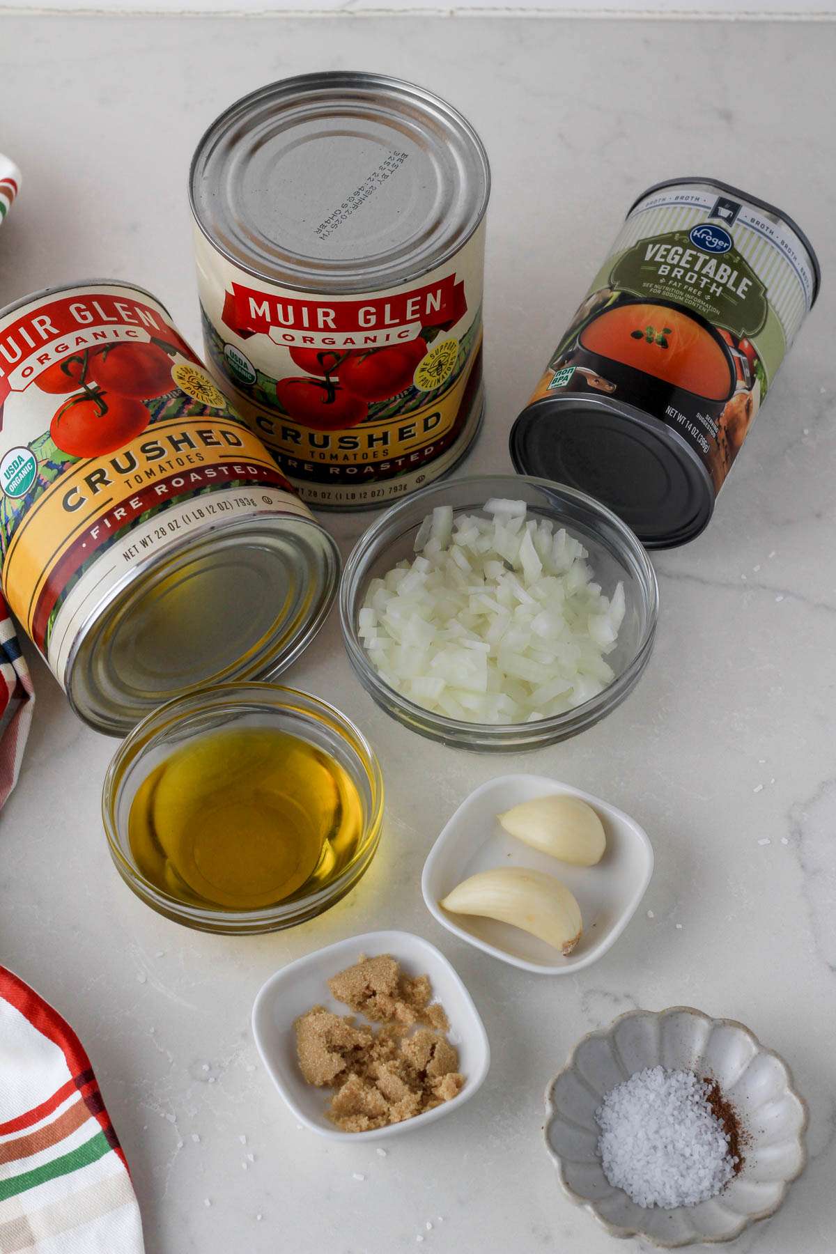 Ingredients for dairy-free tomato soup on a white counter with a dish towel to the left.