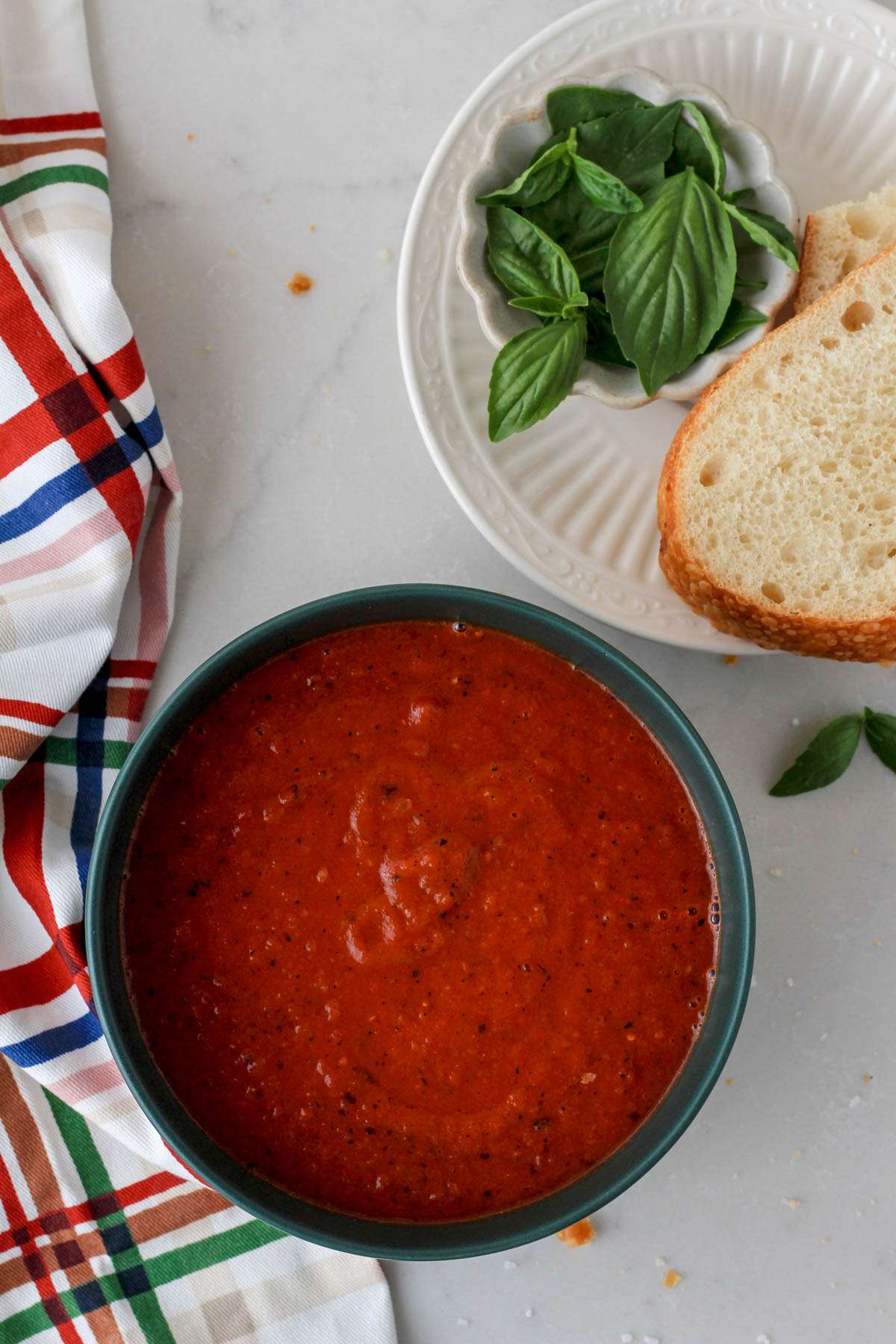A small green bowl filled with dairy-free tomato soup with a small white plate of basil and bread in the top right.