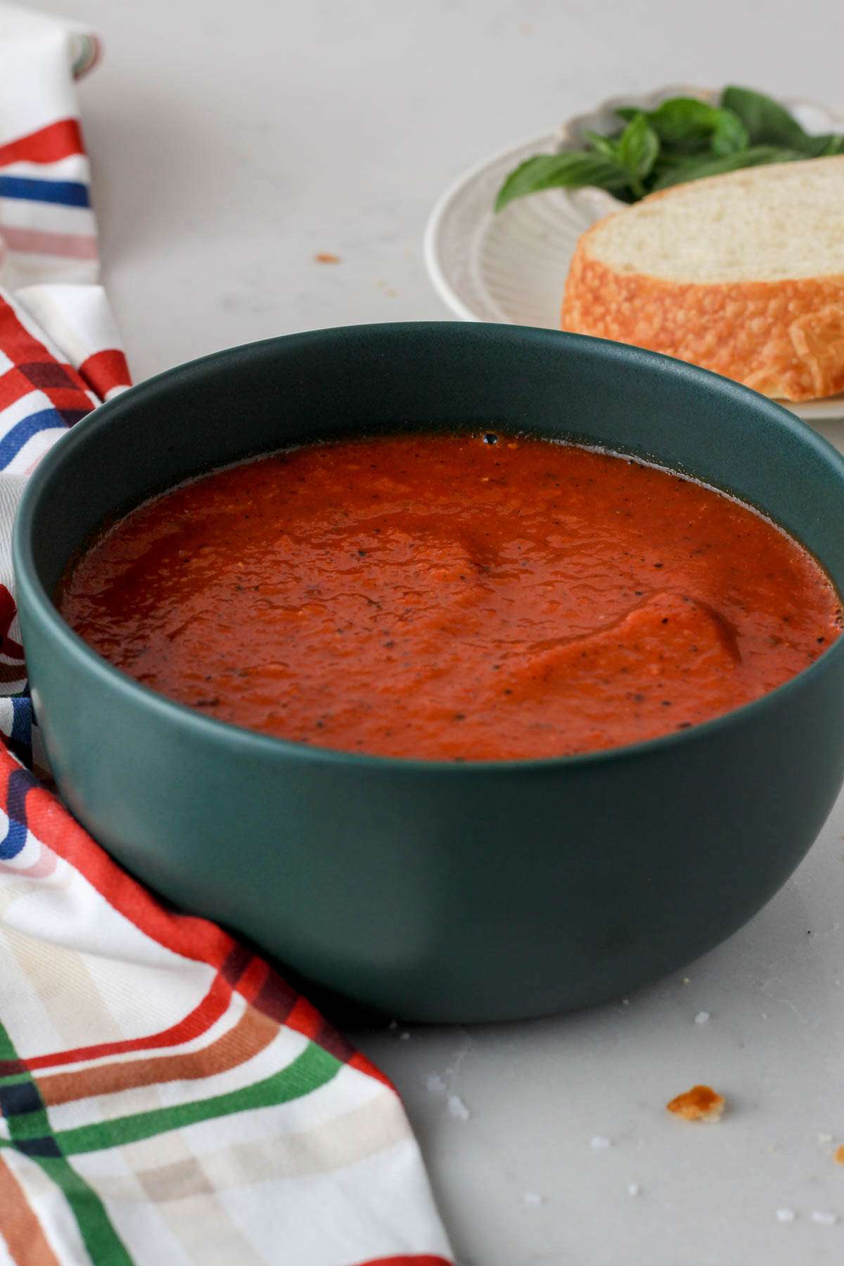 A green bowl filled with gluten-free tomato soup on a white counter in front of a white plate with basil and bread in the back right corner.