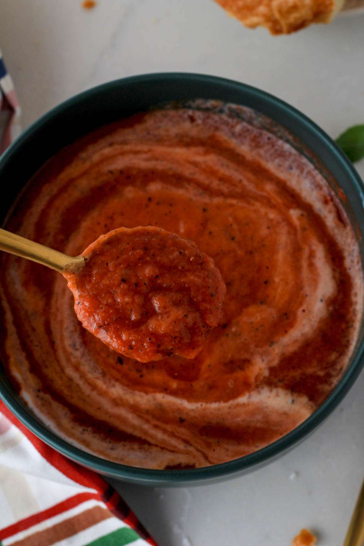 A gold spoon with a scoop of dairy-free tomato soup hovering over a green bowl of tomato soup.