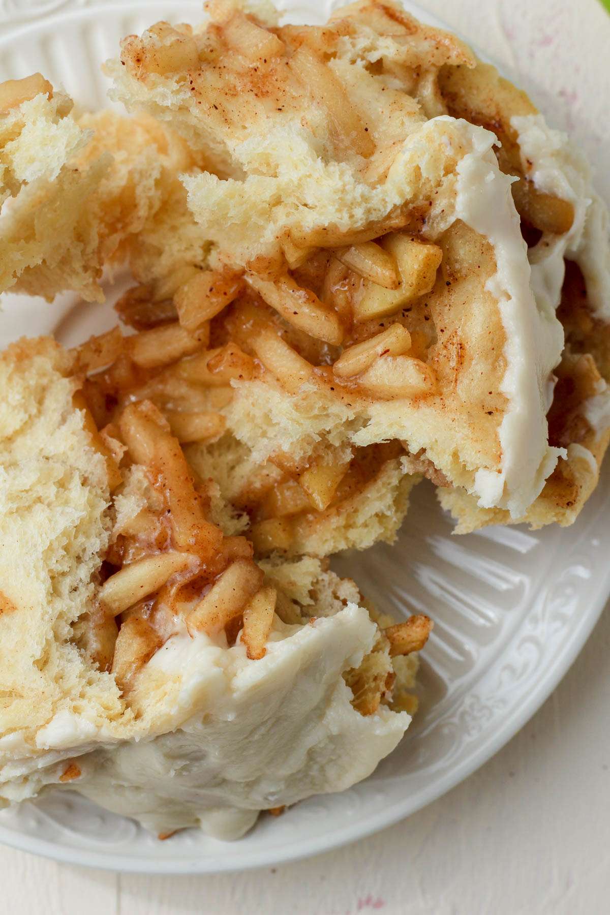 A white plate with a ripped open apple pie cinnamon roll showing the filling.