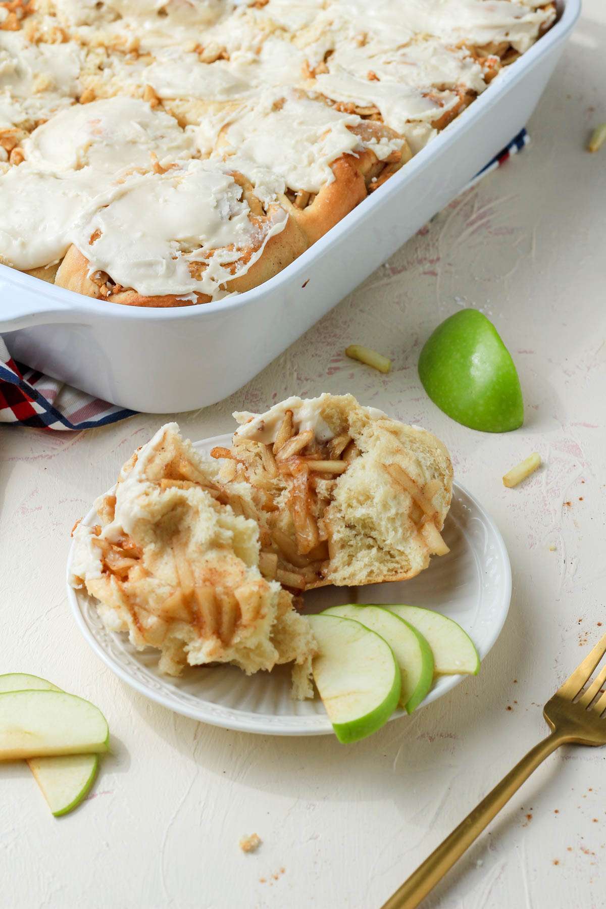 A white plate with a cinnamon roll next to slices of apples with a pan of cinnamon rolls in the back.