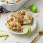 A plate with sliced apples and an apple pie cinnamon roll with a gold fork to the right.
