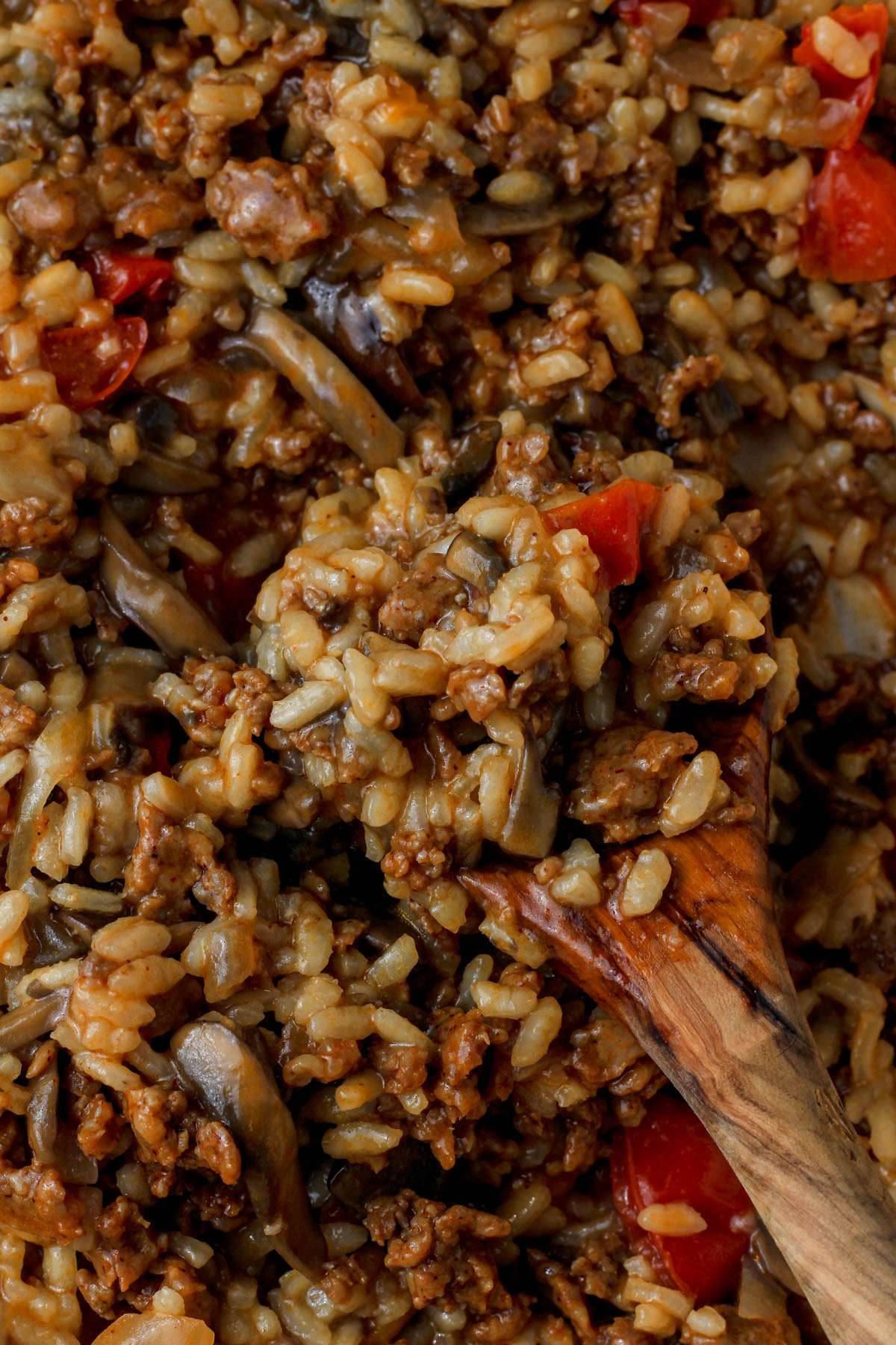 A wooden spoon scooping up chorizo and mushroom risotto with tomatoes in a skillet.