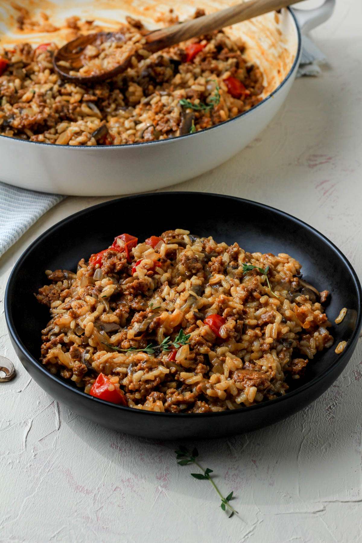 A black bowl of chorizo risotto with mushrooms and tomatoes in front of a white skillet with risotto.