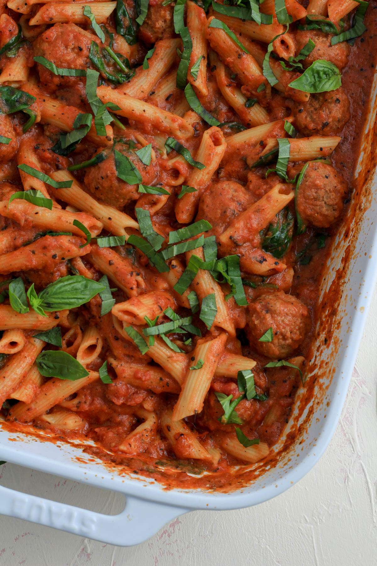 A top down of the front right corner of a white baking dish with dairy free baked penne pasta with meatballs covered in chopped basil.