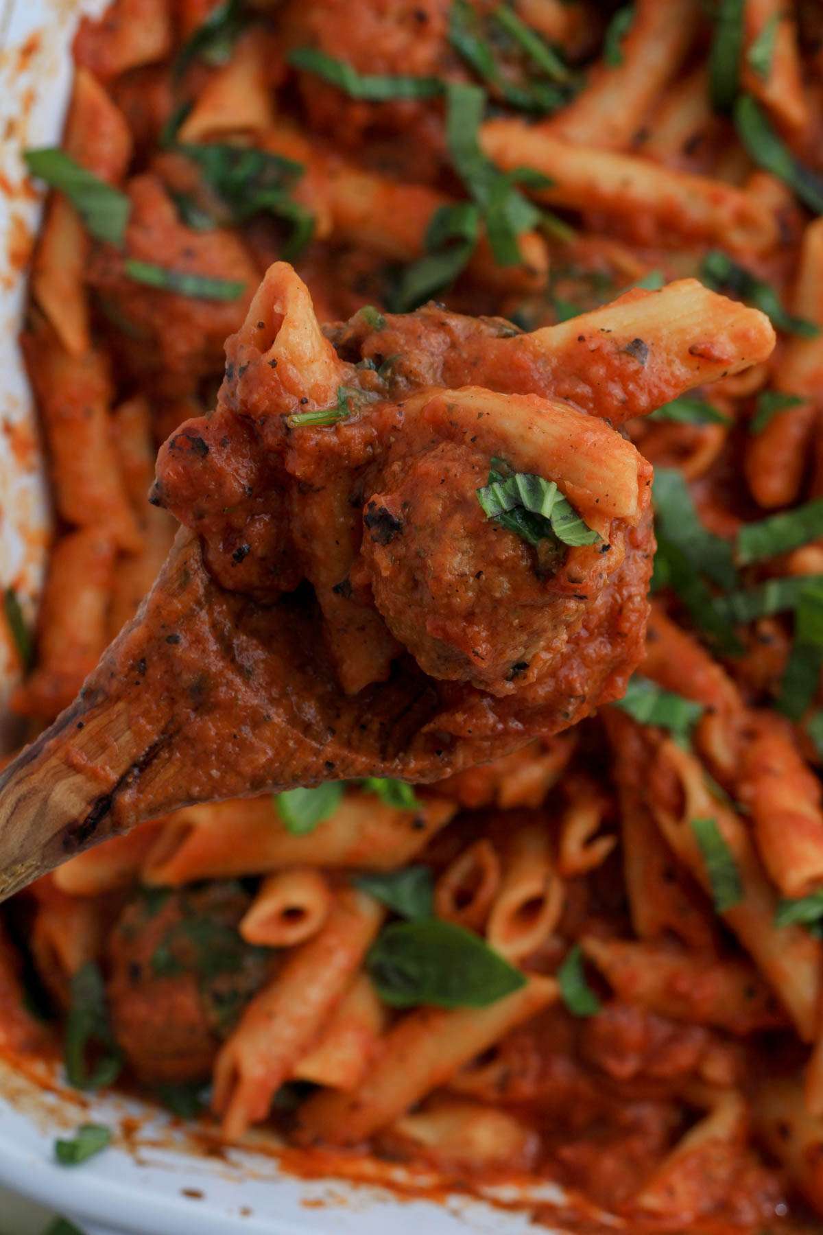 A wooden spoon on the left of the picture holding penne and meatballs over a white baking dish with baked penne.