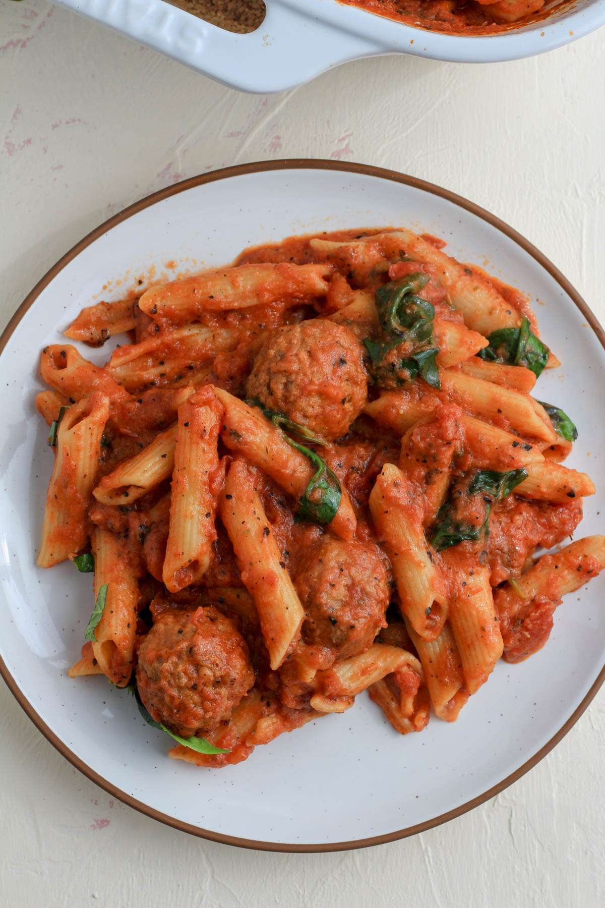 A white plate with a brown rim filled with baked penne and meatballs.