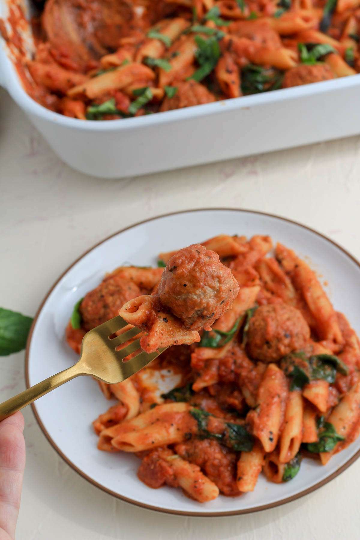 A white plate of dairy free baked penne with meatballs with a fork filled with baked pasta on the left.