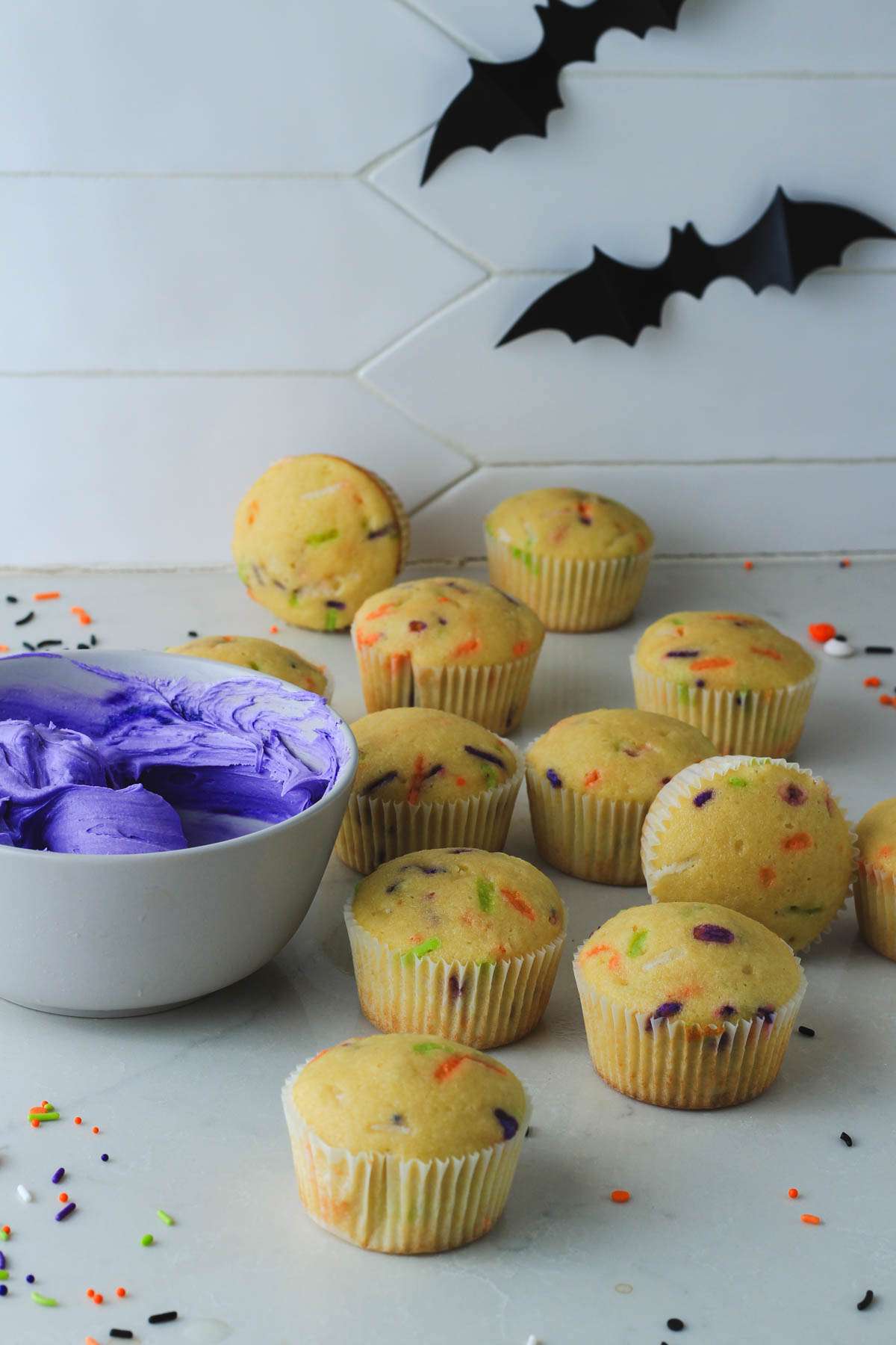 Halloween funfetti cupcakes on a white counter next to a bowl of purple icing.