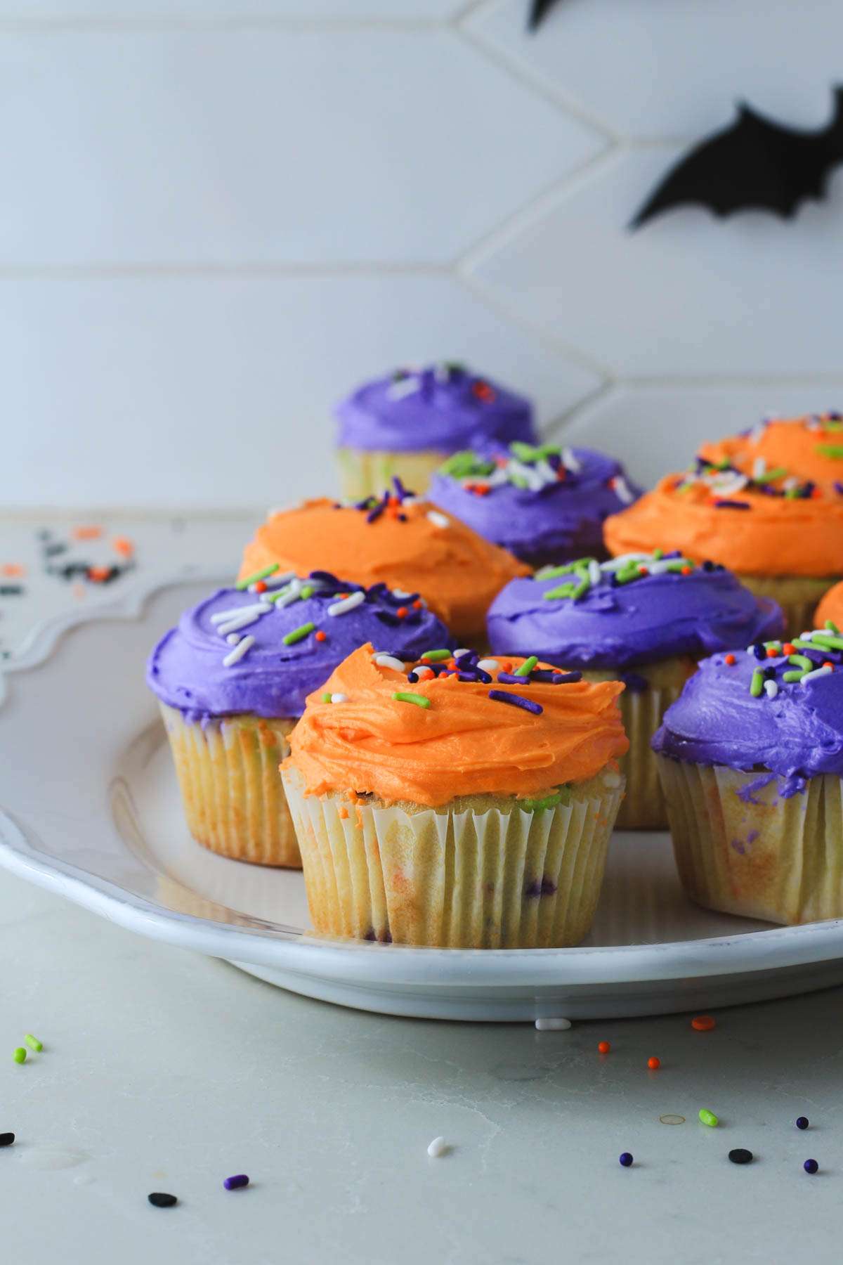 A white platter on a white counter with purple and orange frosted Halloween funfetti cupcakes with a bat in the back right.