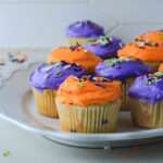 A white platter topped with purple and orange ice Halloween Funfetti Cupcakes.