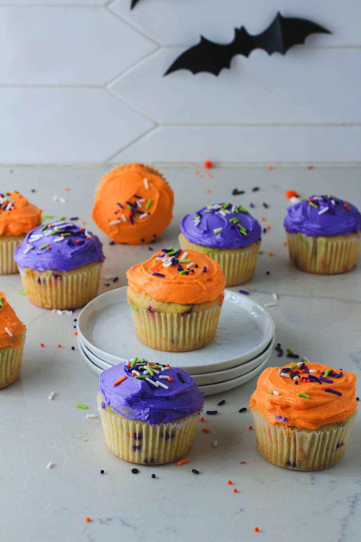 A small plate topped with an orange frosted funfetti cupcake surrounded by orange and purple cupcakes on a white counter.