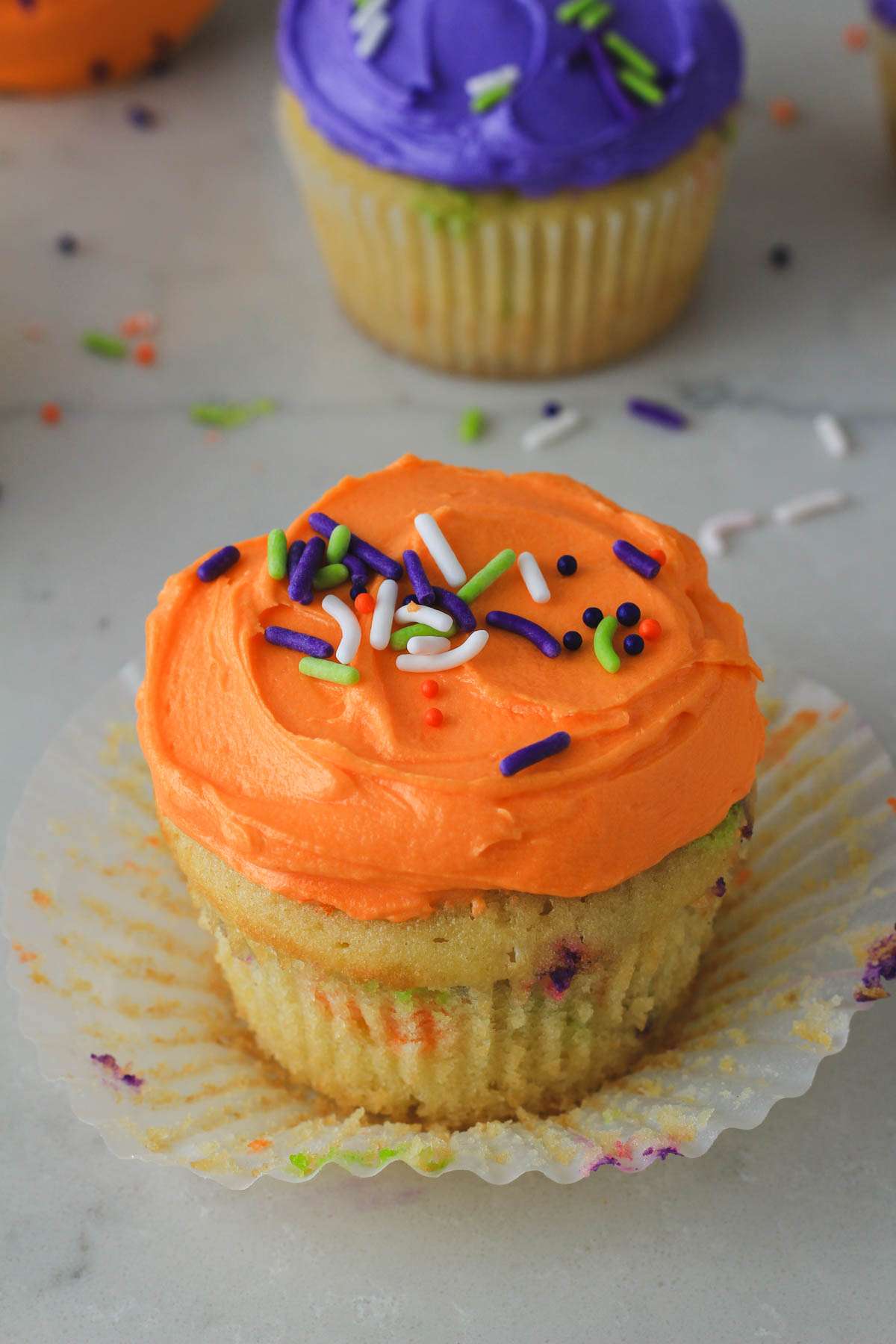 A white counter with an orange frosted Halloween funfetti cupcake with the wrapper pulled off.