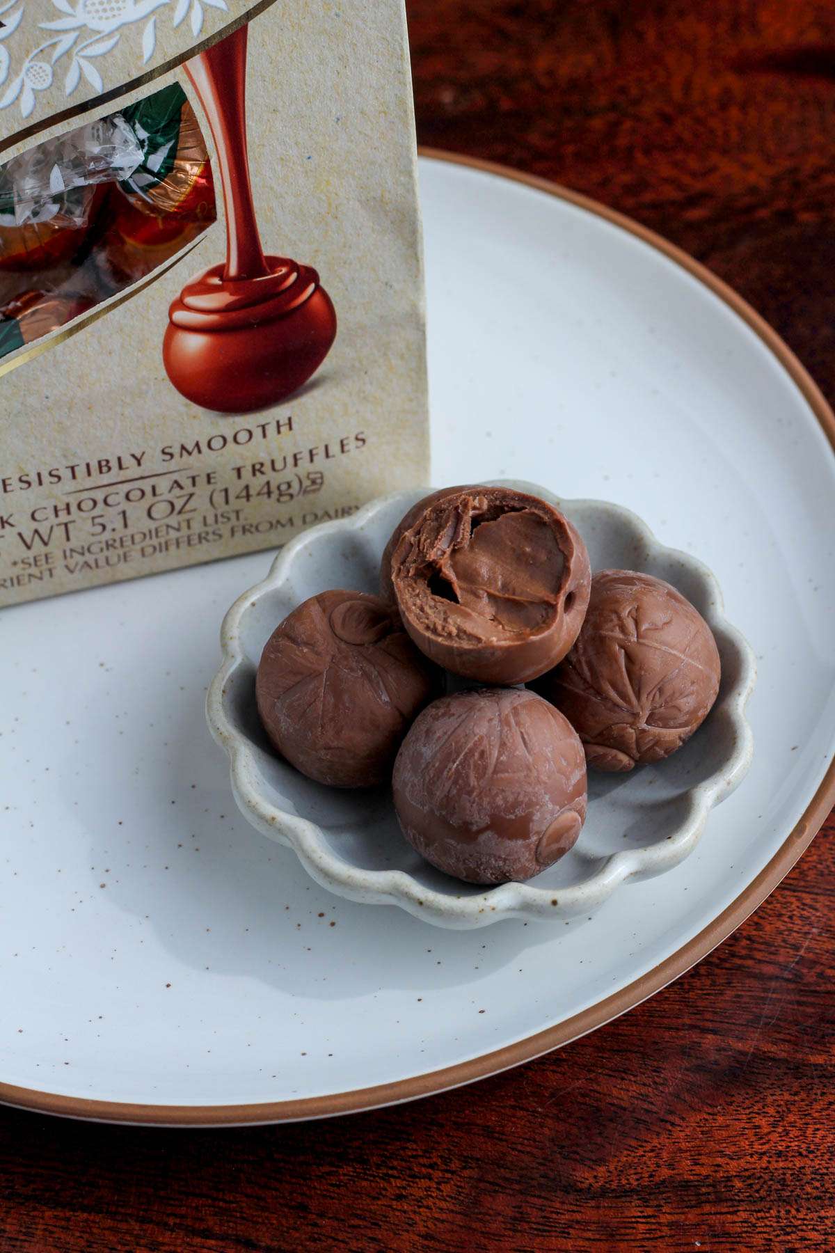 A small bowl with five unwrapped chocolate truffles on a white plate next to a bag of non-dairy Lindt truffles.