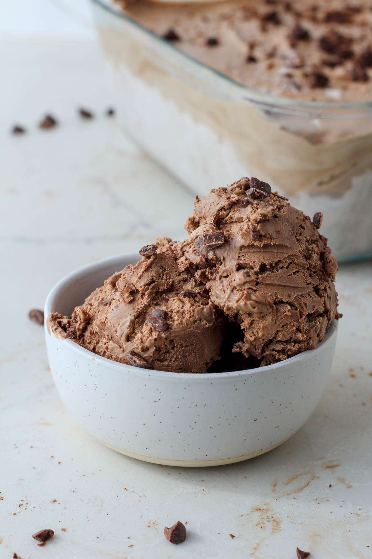 A white bowl in front of a glass pan with scoops of dairy-free chocolate chocolate chip ice cream.