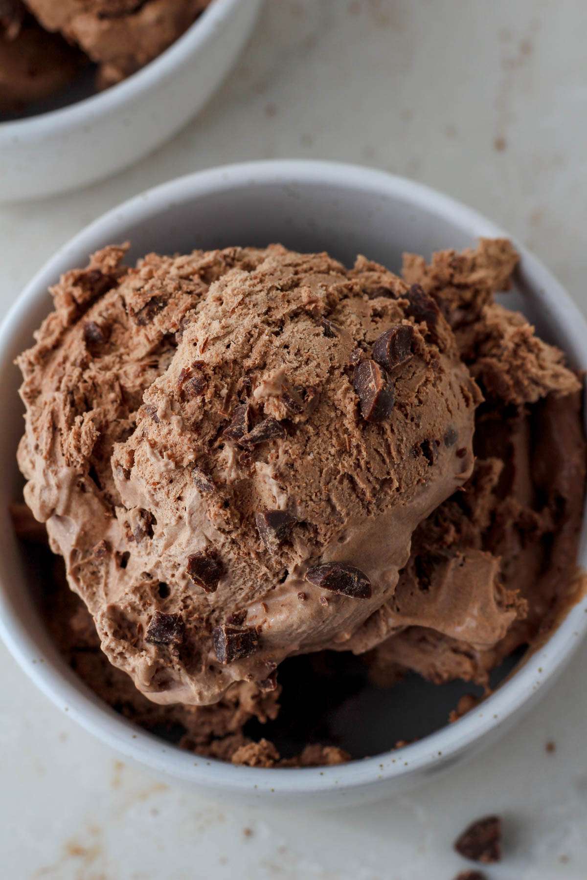 A small white bowl on a white counter with scoops of vegan chocolate ice cream with chocolate chips on top.