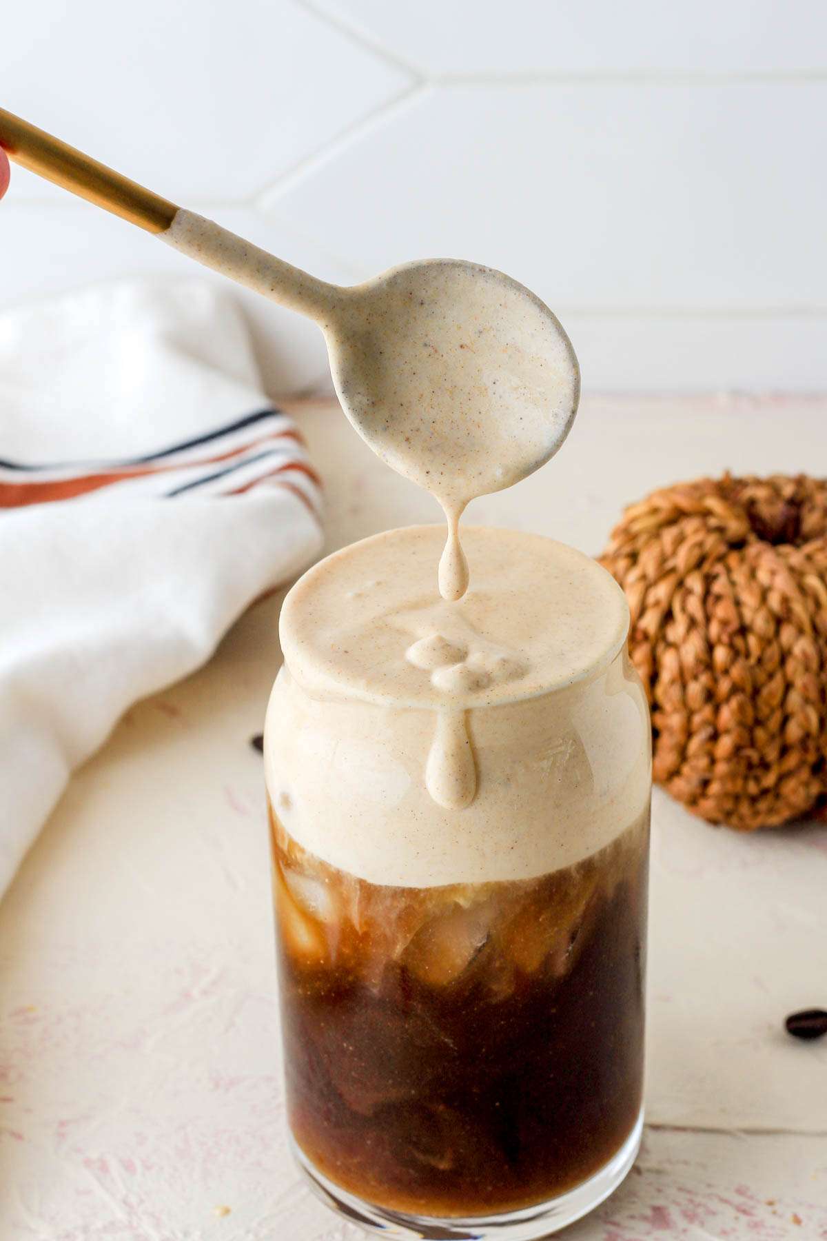 A gold spoon hovering over a glass of ice coffee with pumpkin spice cold foam after mixing the foam into the coffee.