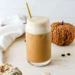 A tall glass of vegan pumpkin cream cold foam topped iced coffee on a cream counter.