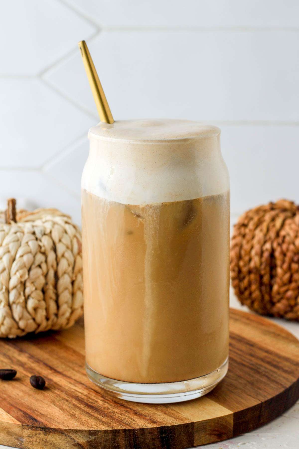 A small wooden plate with a glass of pumpkin spice cold foam topped coffee in front of two woven pumpkins.