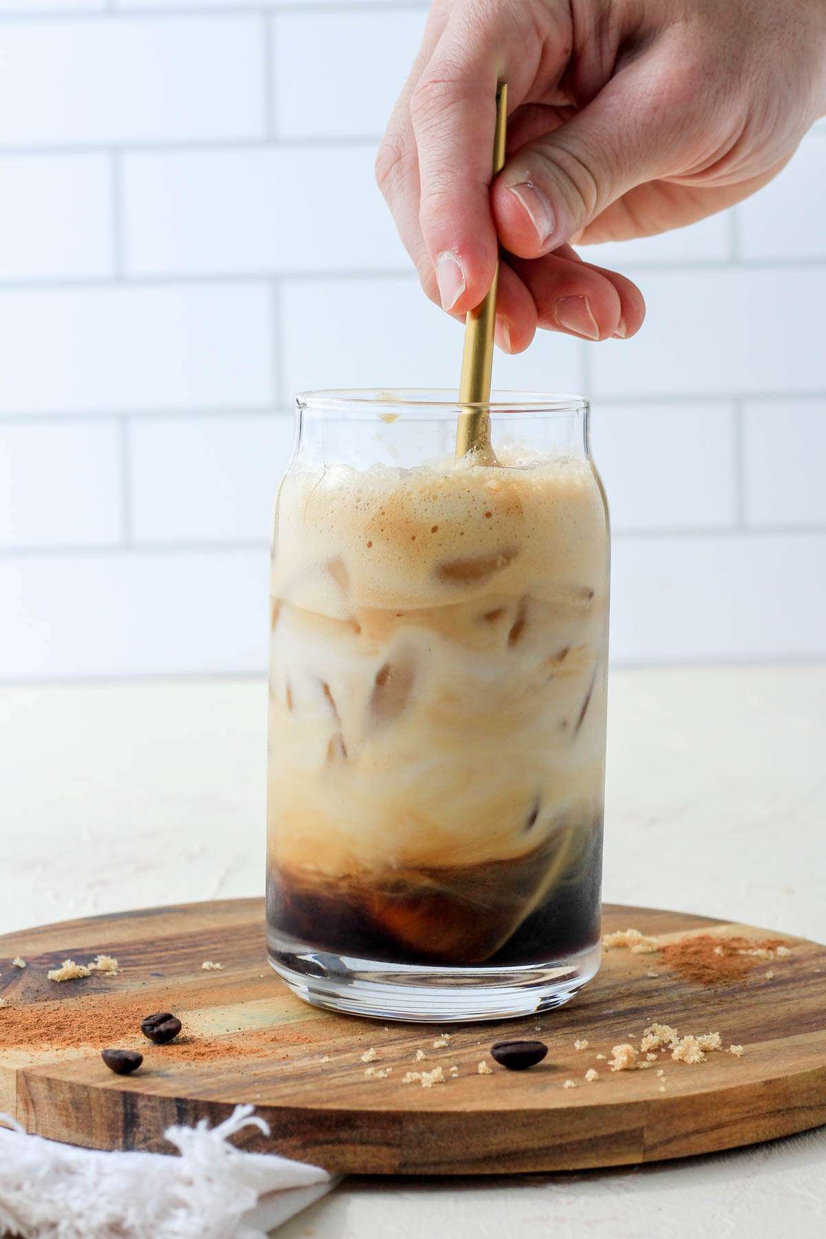 A hand holding a gold spoon mixing the oat milk into the brown sugar shaken espresso.