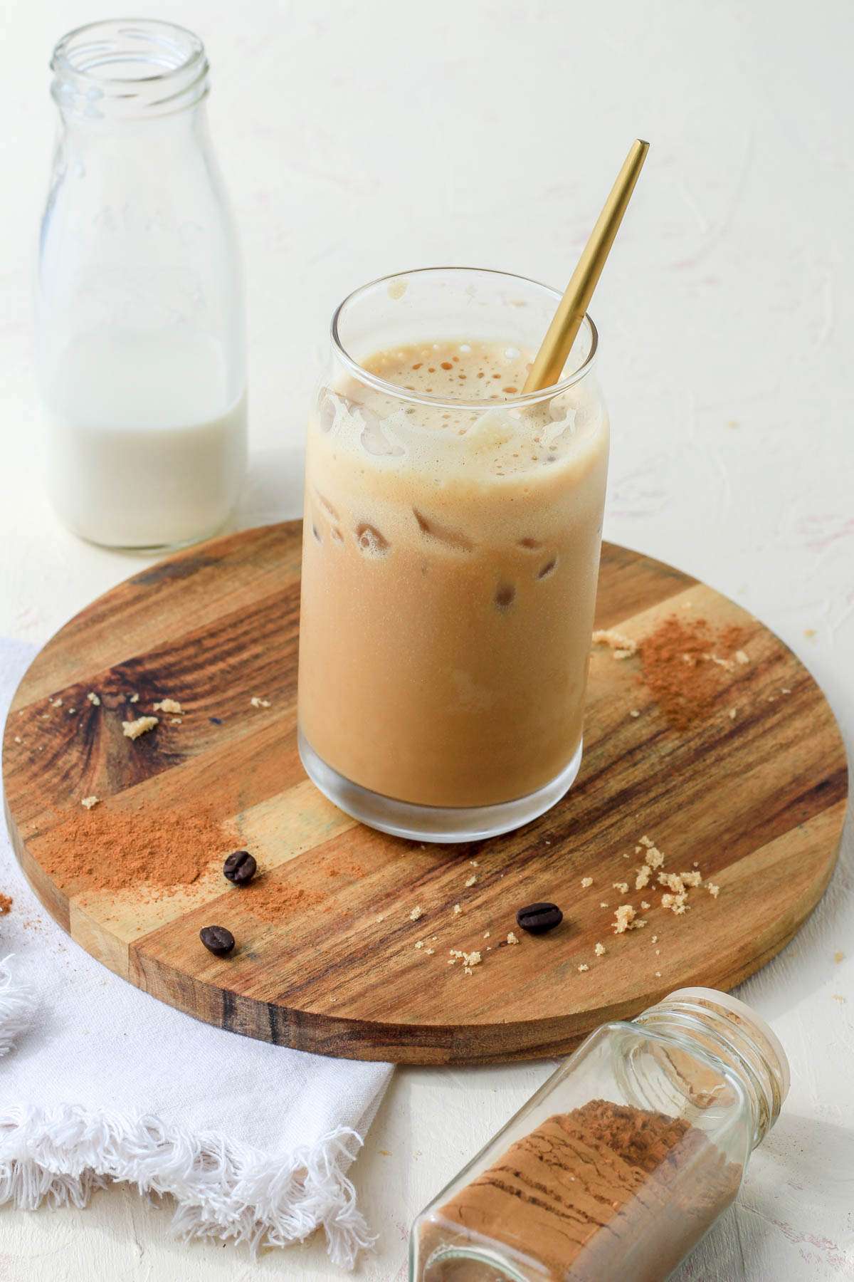 A wooden plate with a brown sugar oat milk shaken espresso in the middle and glass of oat milk in the back left and a jar of cinnamon in the front right.