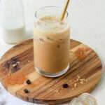 A close up of a brown sugar oat milk shaken espresso on a wooden plate with a jug of oat milk in the back left.