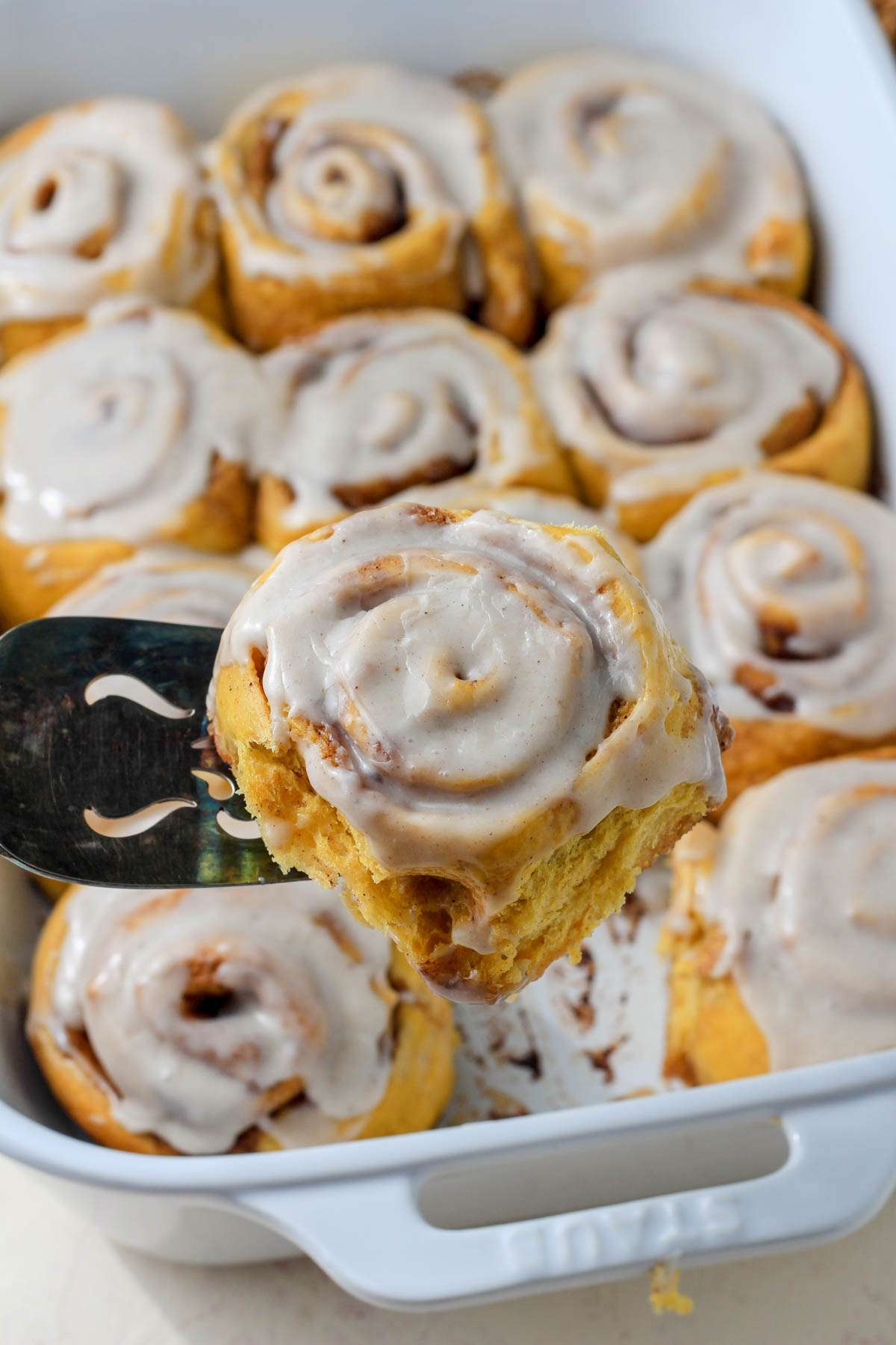 A metal cake server with a dairy-free pumpkin spice cinnamon roll topped with pumpkin spice icing.