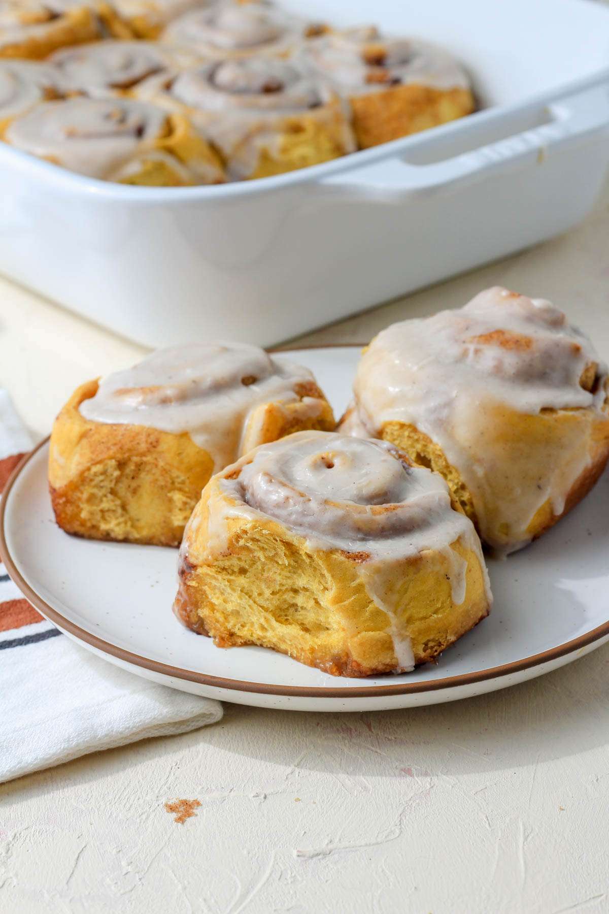 A white plate with a brown rim topped with three pumpkin spice cinnamon rolls in front of a white baking dish of cinnamon rolls.