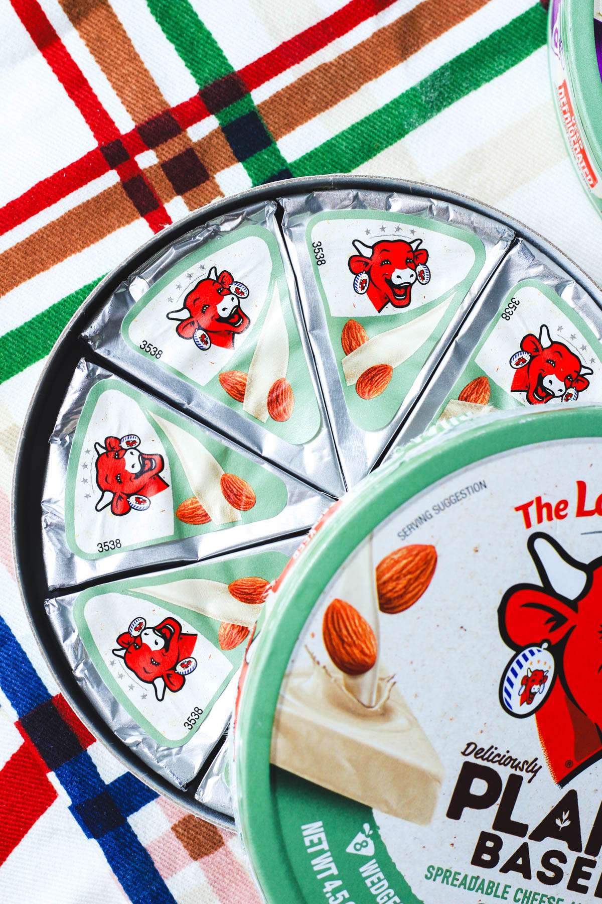 An opened container of laughing cow spreadable plant-based cheese wedges on a colorful dish towel.