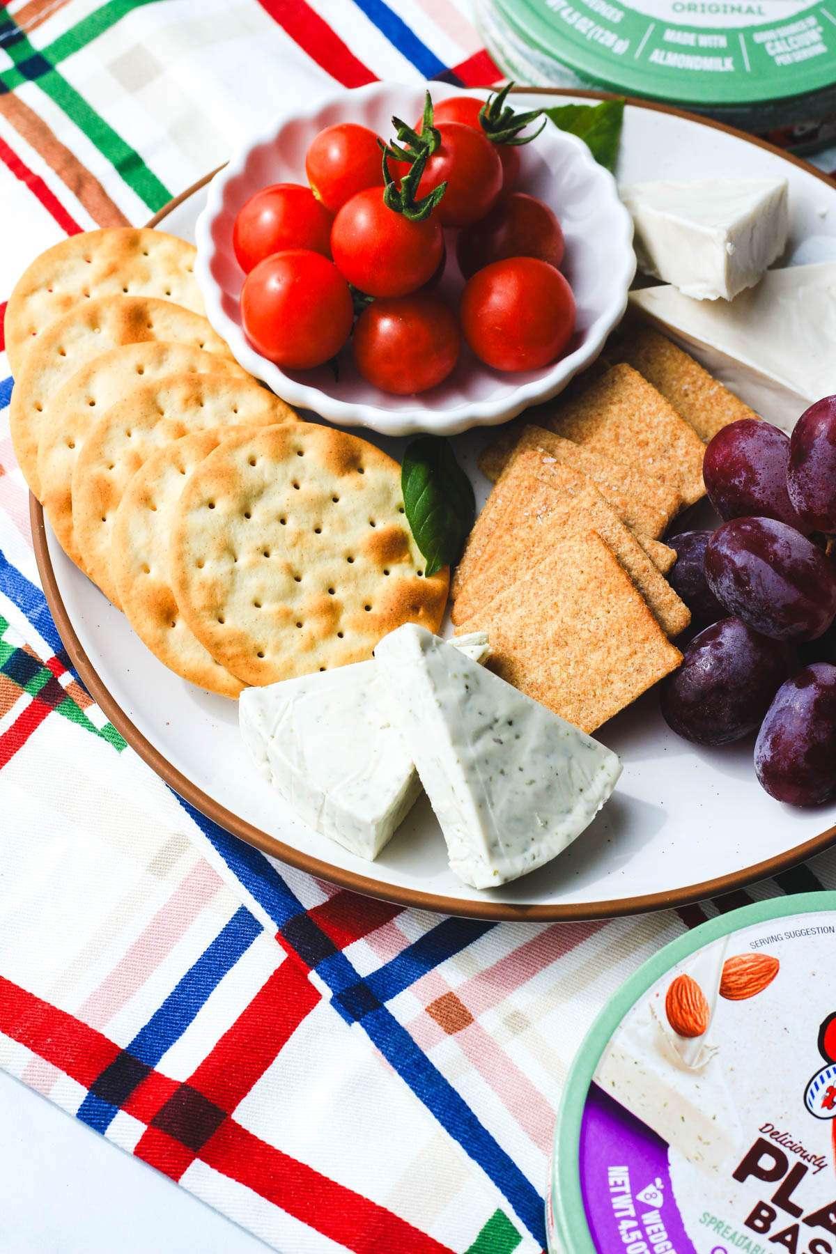 A white plate with a brown rim topped with wedges of plant-based laughing cow cheese wedges, crackers, grapes, and tomatoes.