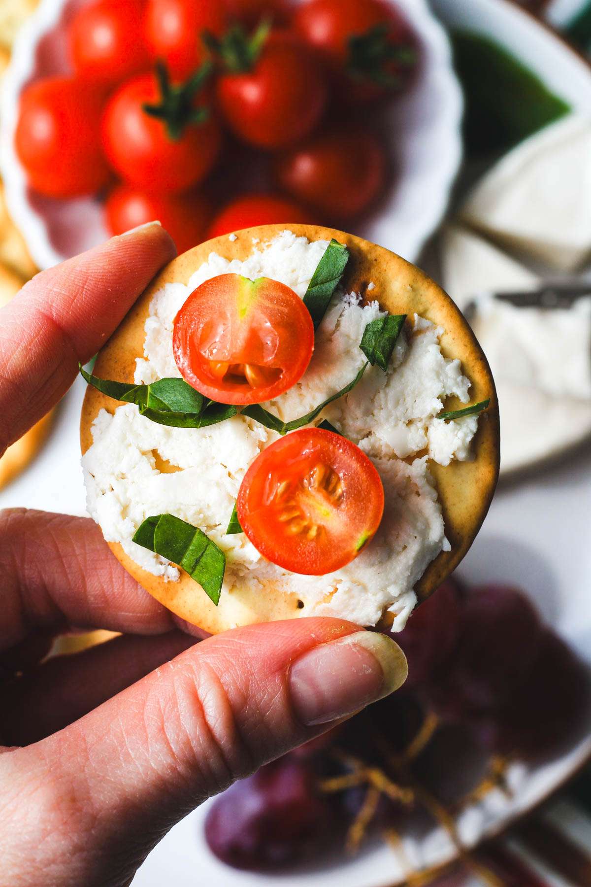 A hand holding a round cracker topped with dairy-free laughing cow cheese, a cut grape tomato, and basil.