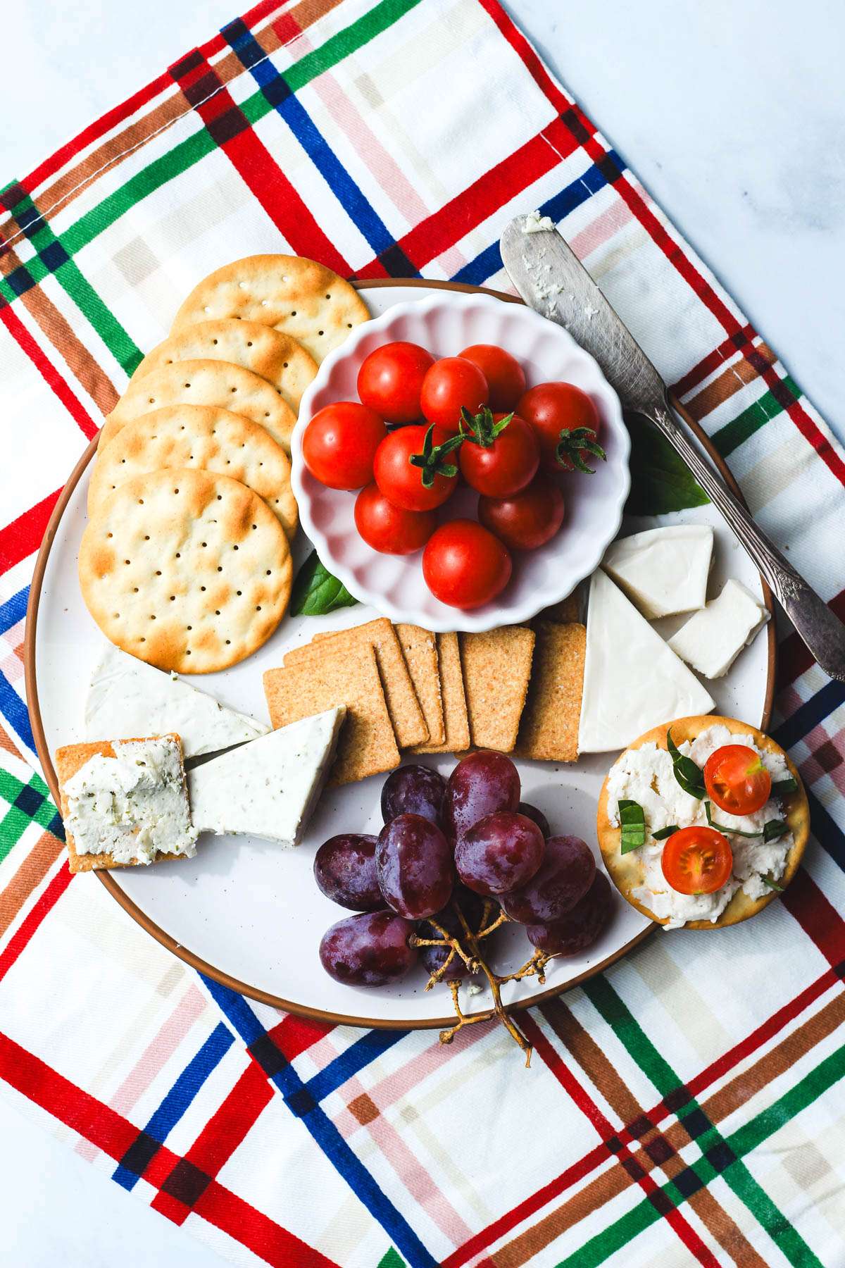 A top down picture of a small snack plate with grapes, tomatoes, crackers, basil, and dairy-free laughing cow cheese wedges.