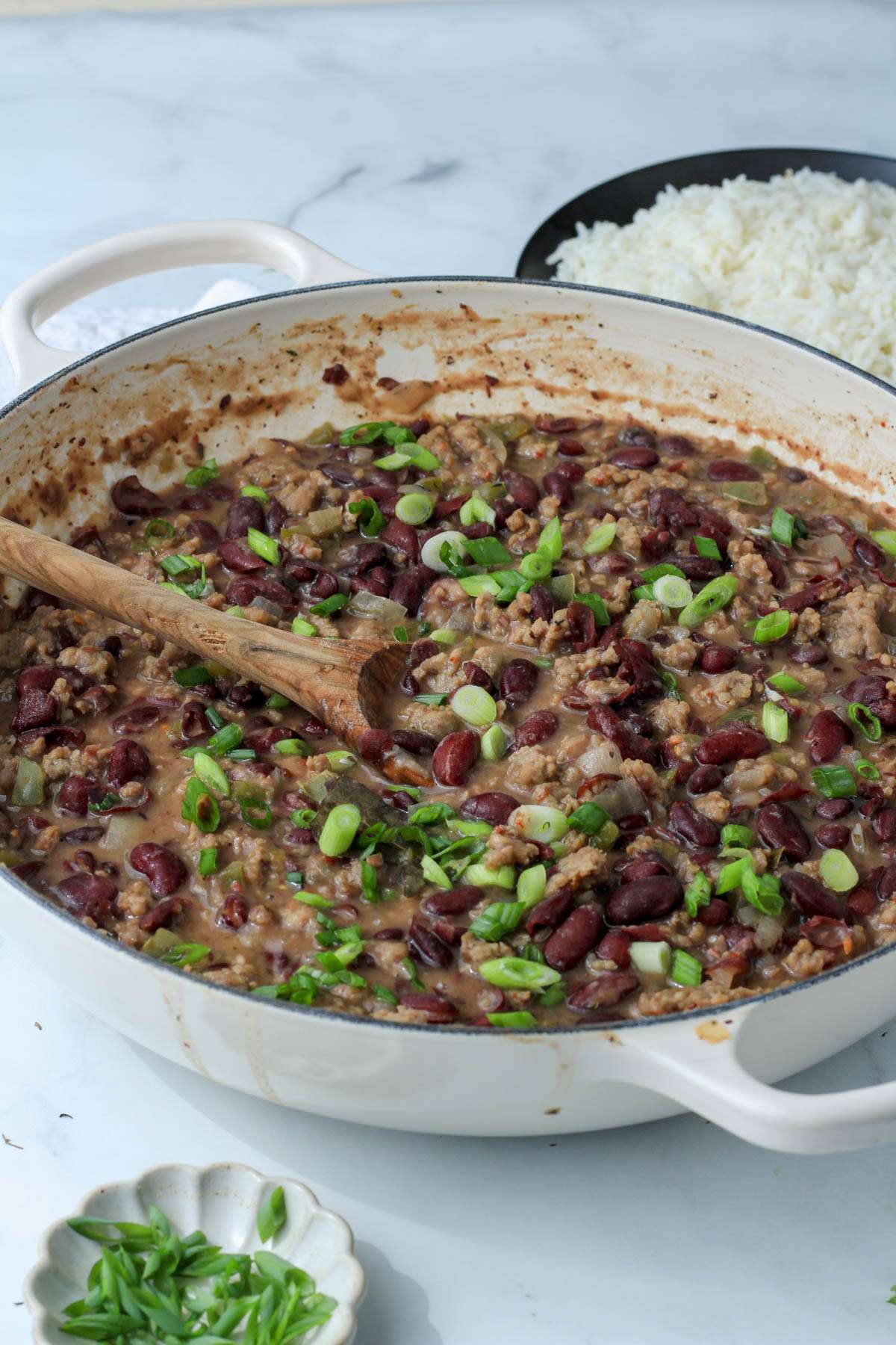 A white pan from the side with a sausage and red bean gravy and a small plate of white rice in the back.