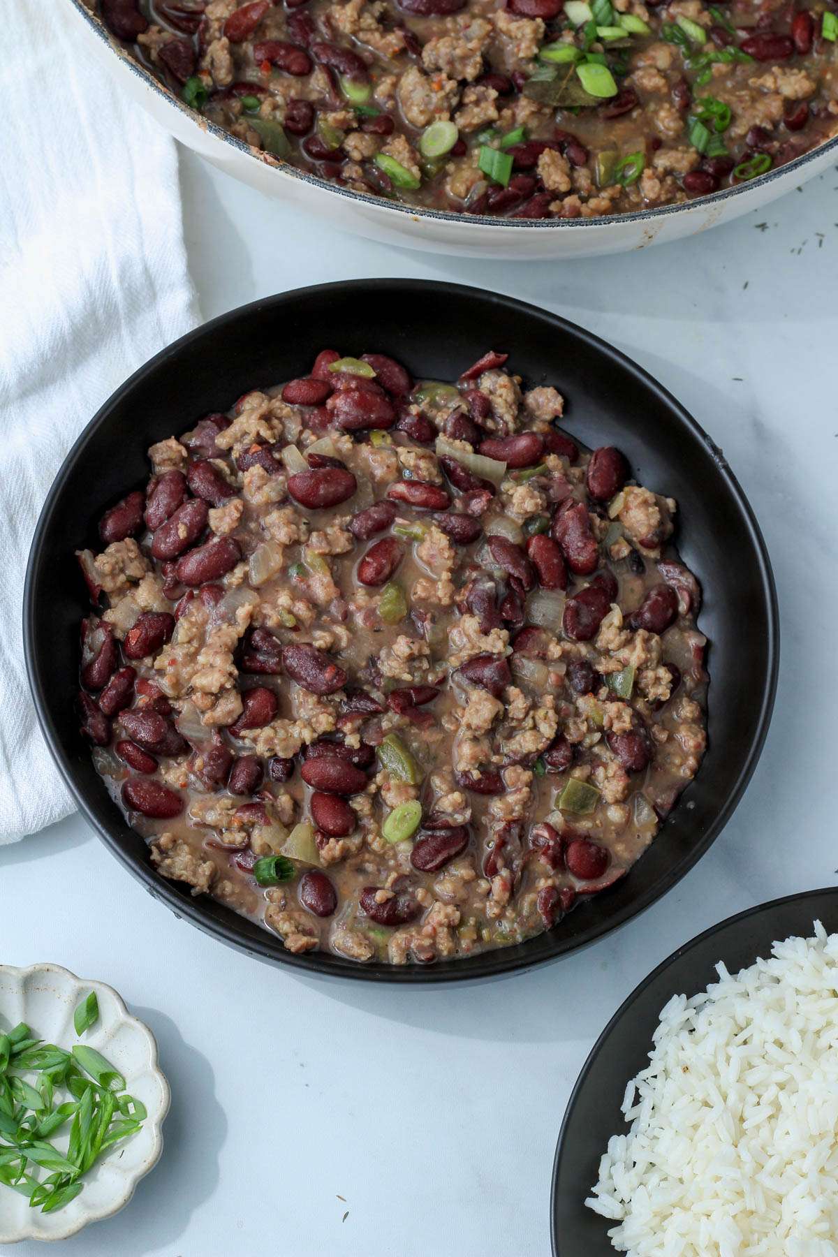 A black bowl filled with red beans and sausage next to a plate of rice and green onion.