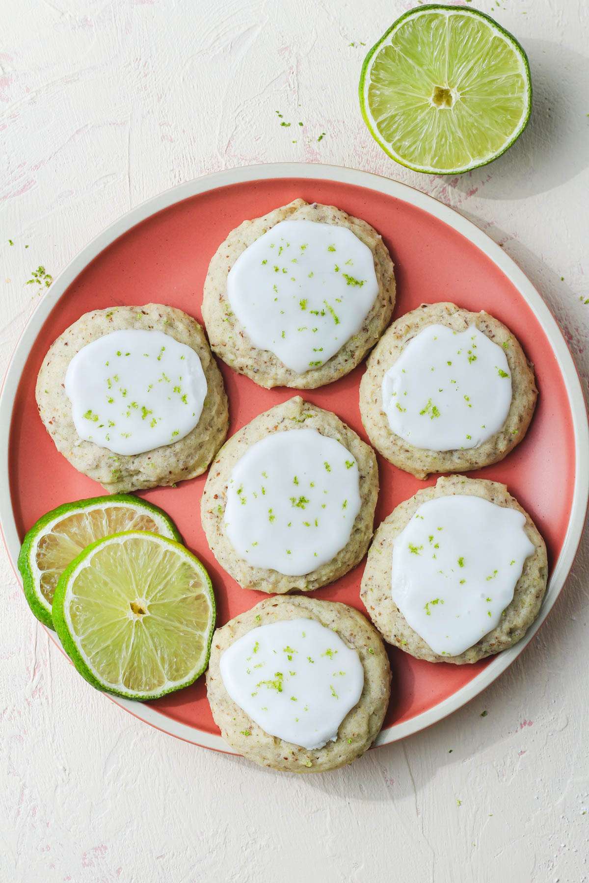 A pink plate with a cream rim topped with vegan lime cookies and two lime slices next to half a lime in the top right corner.