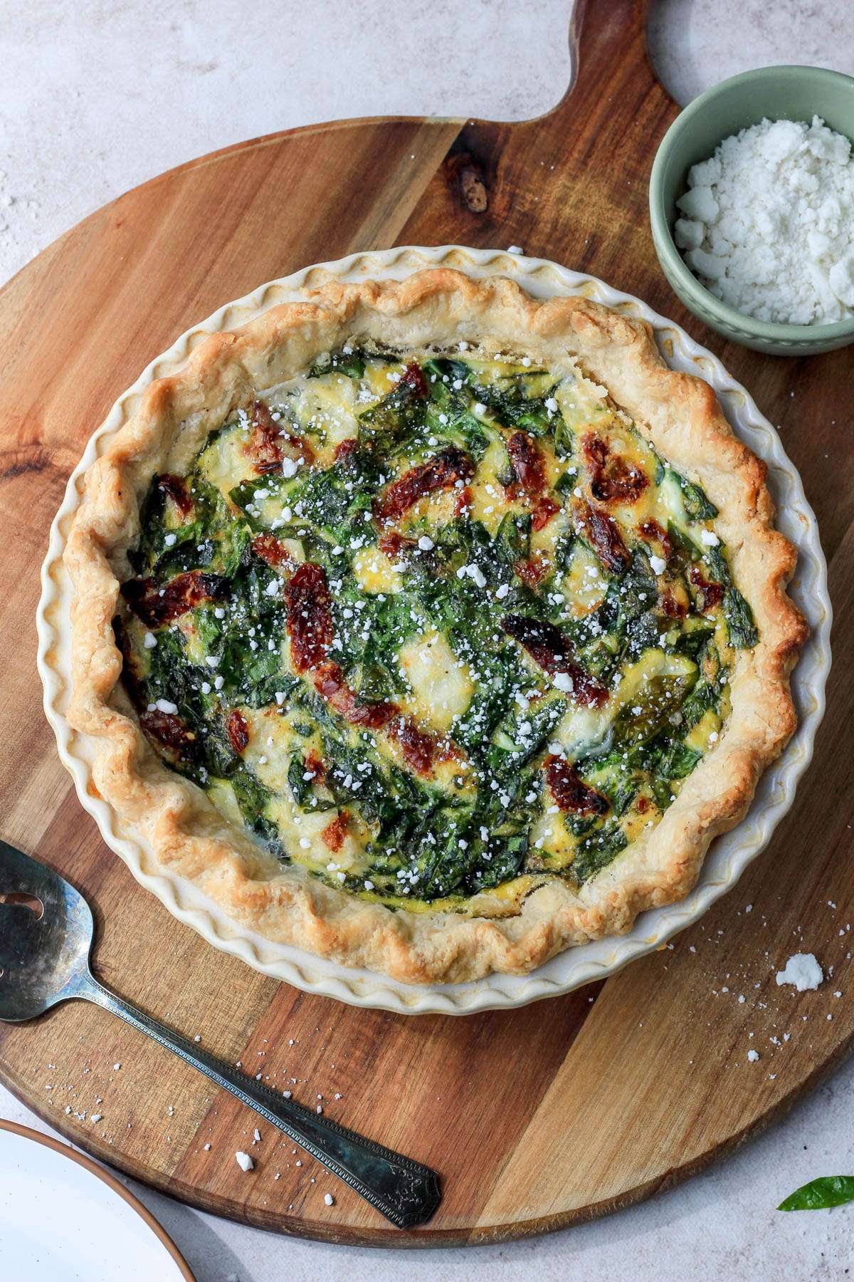 A wooden cutting board with a spinach, feta, and sun dried tomato quiche with a pie server in the bottom left and a small bowl of dairy-free feta in the top right.