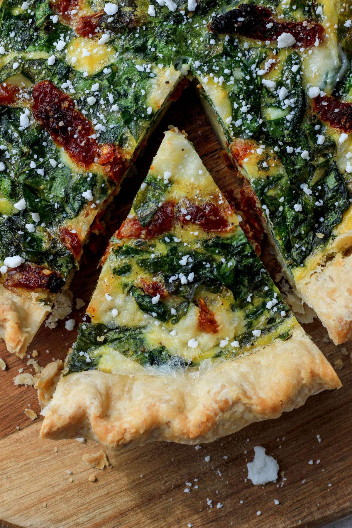 A close up shot of a slice of spinach, feta, and sun dried tomato quiche on a wooden cutting board.