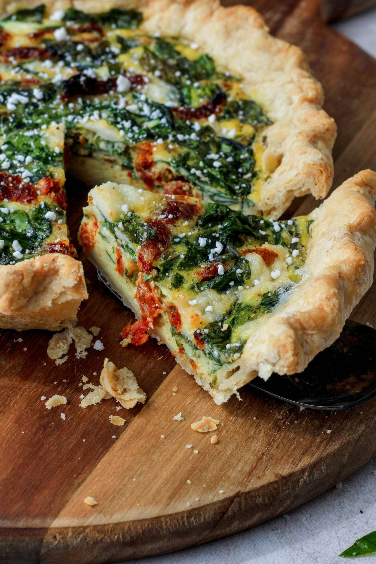 A wooden cutting board with a slice of spinach, feta, and sun dried tomato quiche from the side.