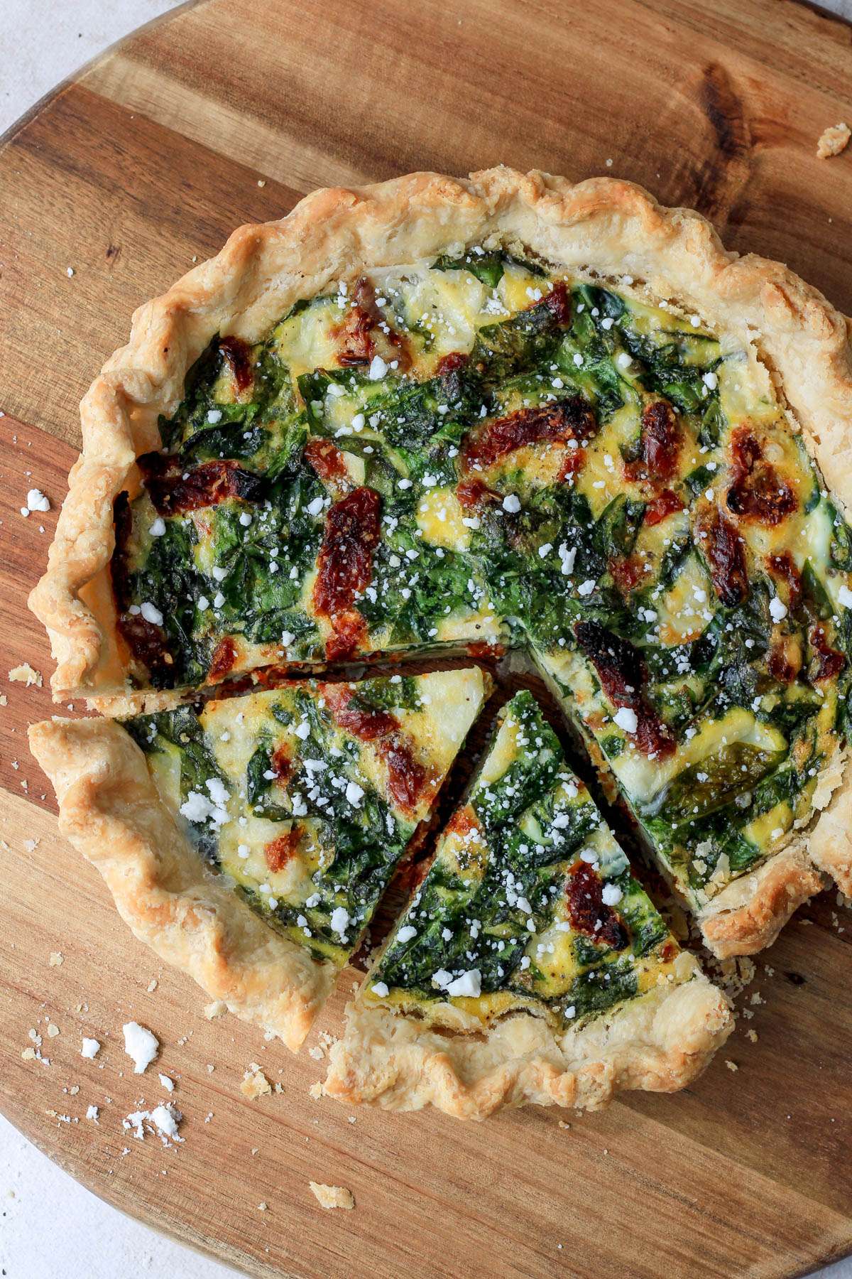A wooden cutting board with a spinach, feta, and sun dried tomato quiche with two slices cut out in the bottom left corner. 