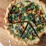 A spinach, feta, and sun dried tomato quiche on a wooden cutting board with two slices cut in the bottom left.
