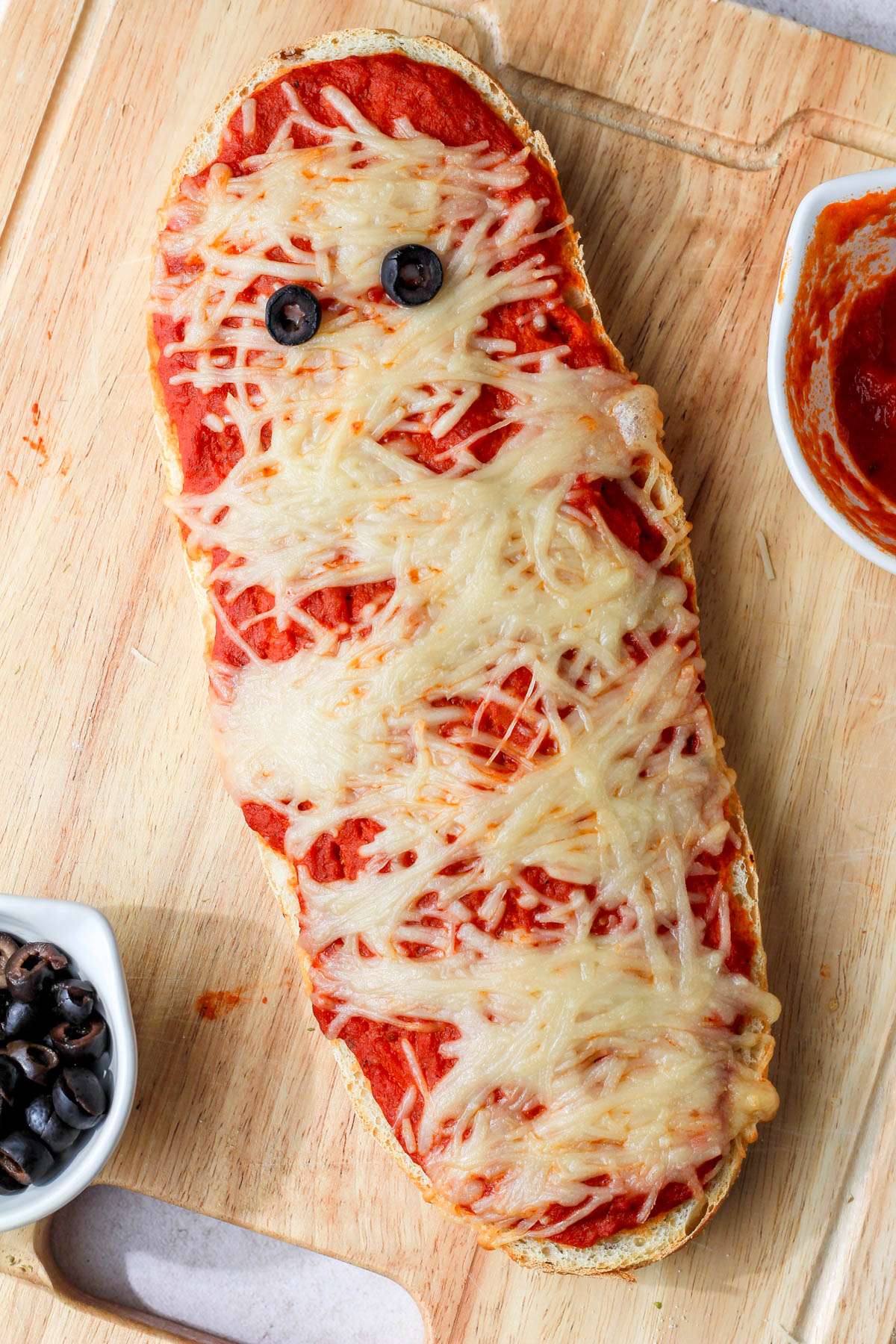 A wooden cutting board with vegan mummy french bread pizza and a bowl of olives and pizza sauce.