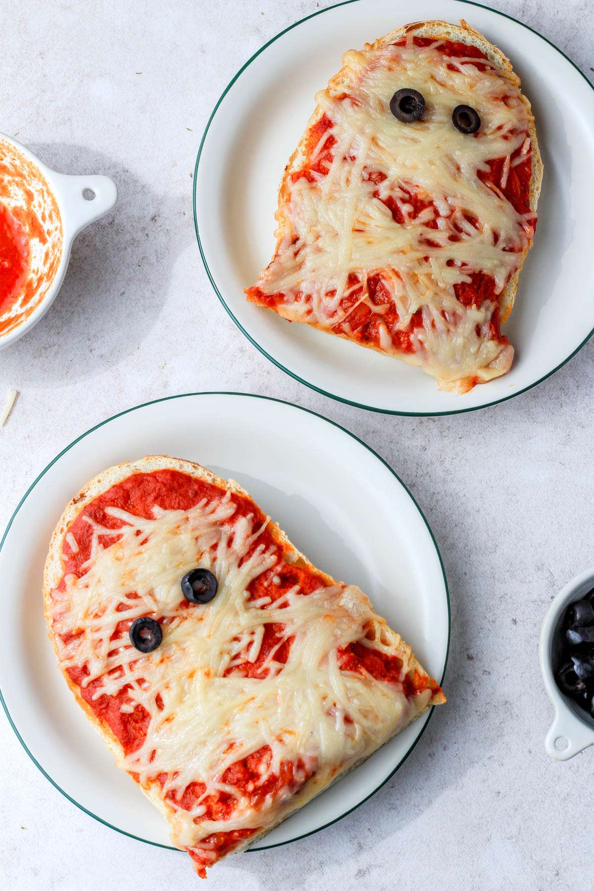 Two white plates with green rims topped with vegan mummy pizza halves.