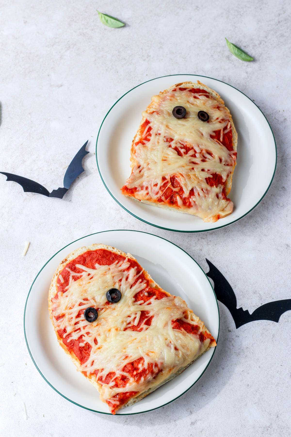 Two plates with half of the vegan mummy pizza on each plate with two black bats on the counter.