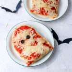 A white plate with a green rim with vegan mummy pizza on french bread with two black bats on the counter.